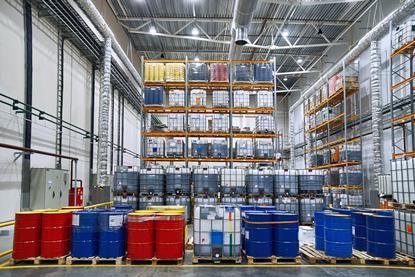 Oil drums and plastic container on pallets in a warehouse on metal shelving.